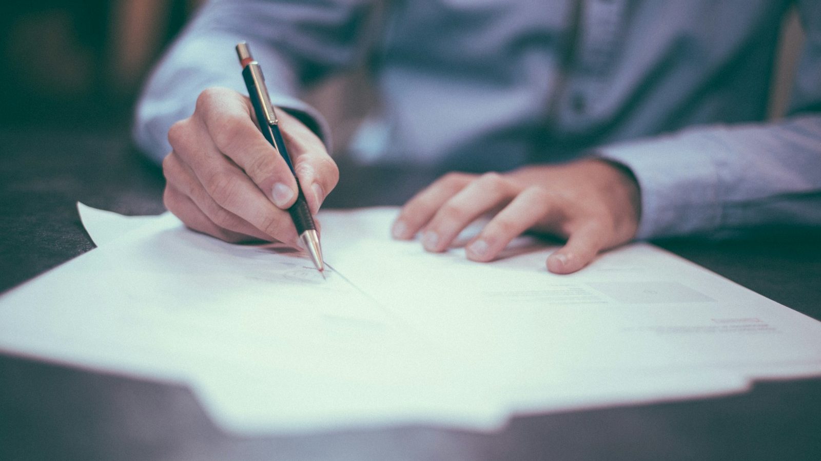 Una mano firma un documento con un bolígrafo sobre una mesa oscura, bajo una luz fría y tenue